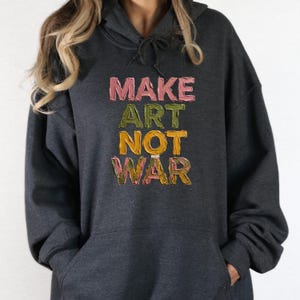 K&ouml;nnte beinhalten: Dunkelgrauer Kapuzenpullover mit dem Text "MAKE ART NOT WAR" in Rosa, Gr&uuml;n und Gelb. Der Kapuzenpullover hat eine Kapuze mit Kordelzug und eine Vordertasche. Der Text ist in einer strukturierten, k&uuml;nstlerischen Schrift.