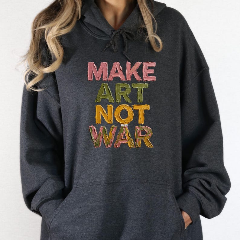 K&ouml;nnte beinhalten: Dunkelgrauer Kapuzenpullover mit dem Text "MAKE ART NOT WAR" in Rosa, Gr&uuml;n und Gelb. Der Kapuzenpullover hat eine Kapuze mit Kordelzug und eine Vordertasche. Der Text ist in einer strukturierten, k&uuml;nstlerischen Schrift.