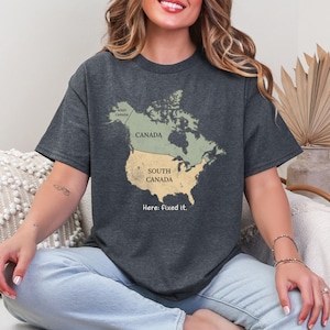 May include: A dark charcoal gray t-shirt featuring a map of North America. The map is divided into two sections: a green top labeled Canada and a yellow bottom labeled South Canada. The text West Canada and Here: fixed it. are also printed.