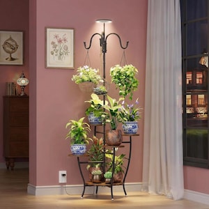 May include: A black metal plant stand with multiple wooden shelves and a built-in LED light. The stand holds various potted plants, including hanging baskets. The pots are decorated with blue and white patterns. The stand is placed in front of a pink wall.