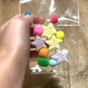 May include: A clear plastic bag containing a collection of small, multi-colored craft supplies, including several fuzzy pom-poms in shades of pink, yellow, orange, green, and blue, alongside multiple star-shaped charms in pale yellow and white.