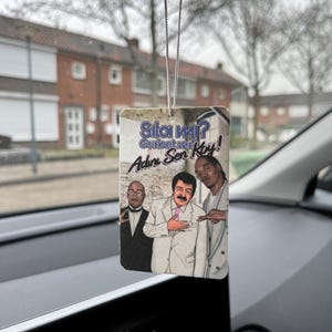 Könnte beinhalten: Rechteckiger Auto-Lufterfrischer mit einem Cartoon-Bild von drei Männern. Der Lufterfrischer hat Text in einer Fremdsprache. Er hängt an einer silbernen Schnur in einem Auto.