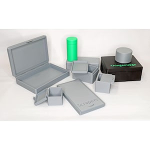 May include: A selection of grey storage boxes and containers in different sizes. There's a rectangular box with an open lid, small square boxes, and a rectangular container with the word "Scrapers" printed on it. A green cylindrical container and a black box with "Thingamajigs" are also visible.