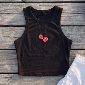 Puede incluir: Un top corto sin mangas de color negro con cuello redondo y un pequeño estampado gráfico de dos frambuesas rojas centrado en el pecho. La prenda cuenta con un acabado acanalado en el cuello y en los bordes de las sisas.