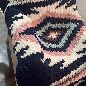 May include: A navy, white, and pink chunky knit blanket with a geometric pattern. The blanket is folded over a light-coloured surface, showcasing its soft texture and colour variations. The design features concentric circles and striped sections.