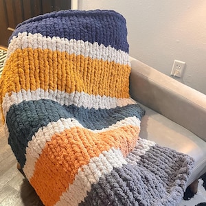 May include: A chunky knit blanket in a striped pattern of navy blue, mustard yellow, white, teal, orange, and gray. The blanket is draped over a gray armchair, showcasing its soft texture and color variations. This cozy throw blanket is perfect for adding warmth and style to any living space.