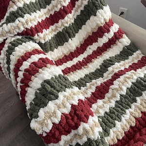 May include: A chunky knit blanket in a striped pattern of red, white, green, and tan. The blanket is folded over a light-colored couch. The thick yarn creates a cozy, textured appearance, perfect for home decor and comfort.
