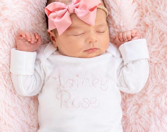 May include: A white long-sleeved baby bodysuit featuring the embroidered name Lainey Rose in light pink script on the chest. The infant wears a matching light pink satin bow headband while lying down with arms raised and eyes closed.