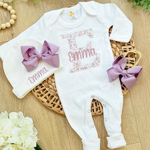 May include: A white baby onesie, hat, and headband set. The onesie features a floral-filled letter "E" and the name "Emma" in pink script. A matching hat and headband with large lavender bows complete the set. The set is displayed on a woven mat.