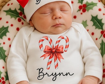 May include: A newborn baby wearing a white onesie and hat, both with the name "Brynn" in script. The hat has a large red bow. The onesie features a candy cane and bow design. The baby is lying in a wicker basket on a Christmas-themed blanket.
