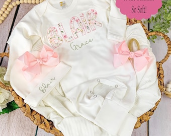 May include: A white long-sleeve baby bodysuit featuring the name BLAIR in a floral print and Grace in green script. Includes a matching white beanie with the name Blair and two light pink bows. Text on the image reads Printed Directly to the Garment So Soft!