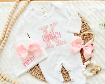 May include: A white long-sleeve baby bodysuit featuring a large letter K with a pink floral pattern and the name Kinsley in pink script. Includes a matching white beanie with a large pink bow and a separate pink bow headband.