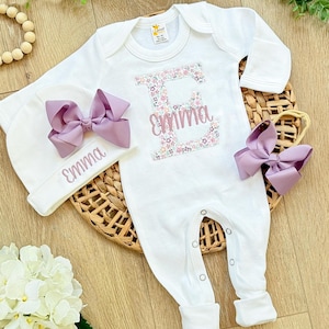 May include: A white long-sleeved baby bodysuit featuring a large letter E with a floral pattern and the name Emma embroidered in pink. The set includes a matching white beanie and a headband, both adorned with large lavender bows and the name Emma.