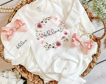 May include: A white long-sleeve baby bodysuit featuring a floral wreath graphic with the name Khloe in gray script. The set includes a matching white beanie with a large pink bow and the name Khloe, plus a separate headband with a pink bow.
