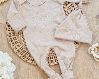 May include: A beige long-sleeved baby bodysuit and a matching knotted beanie hat. Both items feature the name Jack embroidered in white cursive script. The bodysuit has a round neckline and snap closures at the bottom for easy access.