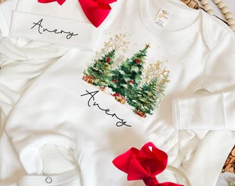 May include: White baby onesie with a Christmas tree design, the name "Avery" is printed on the onesie and matching hat. A red bow is attached to the hat and another red bow is on a headband. The onesie has long sleeves.
