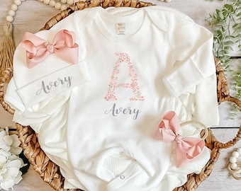 May include: A white long-sleeve baby bodysuit and matching beanie set. The bodysuit features a large floral letter A and the name Avery in gray script. The beanie is also personalized with the name Avery. Both items include a dusty pink bow.