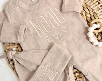 May include: A long-sleeved beige baby bodysuit and matching knotted hat set. The bodysuit features the embroidered name Noah Hayes in white thread. The hat features the embroidered name Noah in white thread. Both items have a soft, heathered texture.