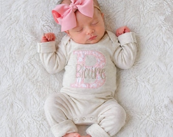 May include: A sleeping infant wearing a long-sleeved, heathered beige bodysuit featuring a large pink patterned letter B with the name Blaire embroidered in script across the center. A large, solid pink hair bow is attached to a thin headband.