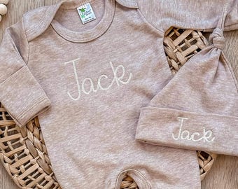 May include: A long-sleeved heather beige baby bodysuit and a matching knotted beanie hat. Both items feature the name Jack embroidered in white thread. The bodysuit has a round neckline and snap closures at the bottom for easy access.