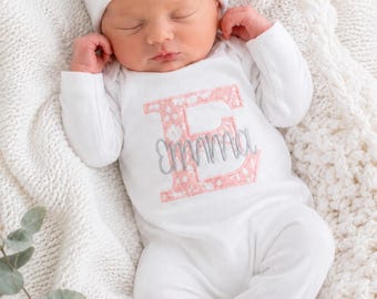 May include: A white long-sleeved baby bodysuit and matching beanie hat. The beanie features a coral pink bow and the name Emma embroidered in gray. The bodysuit displays a large coral floral letter E with the name Emma embroidered in gray.