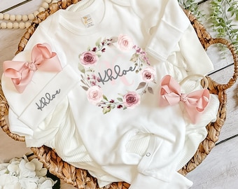 May include: A white long-sleeve baby bodysuit featuring a floral wreath graphic with the name Khloe printed in the center. The set includes a matching white baby hat with a large pink bow and the name Khloe, plus a separate pink hair bow.