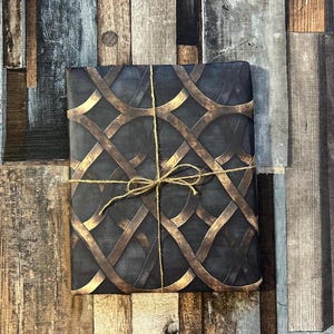 May include: A present wrapped in dark blue paper with a gold geometric design. The gift is tied with twine and a bow. The background is a wooden surface with shades of brown, grey, and blue.