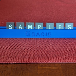 Puede incluir: Un soporte de palabras de plástico azul con la palabra "SAMPLES" escrita en baldosas grises con letras blancas. El nombre "Gracie" está impreso en la parte delantera. El soporte está sobre una superficie roja.
