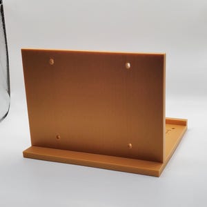 Puede incluir: Un expositor rectangular de color marrón claro con una parte trasera vertical y una base horizontal. El expositor tiene dos pequeños agujeros cerca de la parte superior y está diseñado para sujetar objetos. El expositor está hecho de un material sólido.