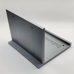 Könnte beinhalten: Eine graue, L-förmige Halterung mit strukturierter Oberfläche. Die Halterung hat drei Löcher und das Wort "Inzane" ist auf die horizontale Oberfläche graviert. Die Halterung besteht aus einem festen Material.