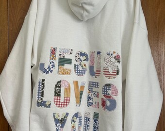 May include: White hooded sweatshirt with the phrase "JESUS LOVES YOU" appliqued on the back. The letters are made of various floral and patterned fabrics. The sweatshirt has a drawstring hood and ribbed cuffs.