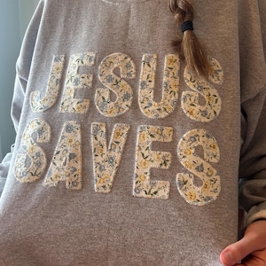 Könnte beinhalten: Graues Sweatshirt mit dem aufgenähten Schriftzug "JESUS SAVES" auf der Vorderseite. Die Buchstaben bestehen aus einem floralen Stoff mit blauen und gelben Blumen. Das Sweatshirt hat einen Rundhalsausschnitt.