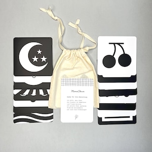 Könnte beinhalten: Ein Set schwarz-weißer Lernkarten mit verschiedenen grafischen Designs, darunter ein Mond und Kirschen, zusammen mit einem kleinen Kordelzugbeutel. Der Beutel enthält eine Karte mit deutschem Text. Die Karten sind für Bildungszwecke konzipiert.