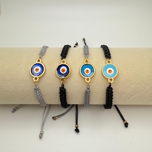 May include: Four adjustable bracelets with gold-colored circular charms featuring an evil eye design. The evil eye charms have blue, orange, and white details. The bracelets have gray and black braided cords.