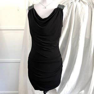 May include: A black sleeveless dress featuring a cowl neck and ruched detailing. The shoulders are embellished with beads. The dress is displayed on a mannequin against a white backdrop, highlighting its elegant design.