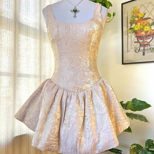 Może przedstawiać: Jasnoróżowa, teksturowana sukienka z kwadratowym dekoltem i rozkloszowaną spódnicą. Sukienka jest prezentowana na manekinie. Sukienka ma dopasowany gorset i krótką, plisowaną spódnicę. Tkanina ma subtelny połysk, nadając jej wygląd vintage.