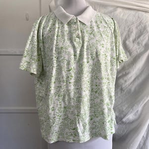 May include: White polo shirt with a green floral pattern. The shirt has short sleeves and a white collar. The floral design features delicate green outlines of flowers and leaves, creating a soft, all-over pattern. The shirt is buttoned at the neckline.