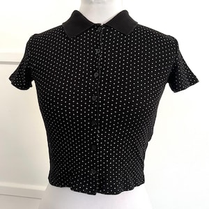 May include: Black short-sleeved polo shirt with white polka dots. The shirt has a black collar and a button-down front. The fabric appears to be a knit material. The shirt is displayed on a white mannequin.