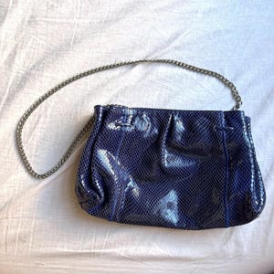 May include: A navy blue handbag with a silver chain strap. The bag has a textured, snakeskin-like pattern. The bag is rectangular with a gathered top.