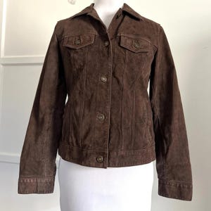 May include: A brown suede jacket with a collared neckline, two chest pockets, and button closures. The jacket has long sleeves and a tailored fit. The color is a rich, dark brown.
