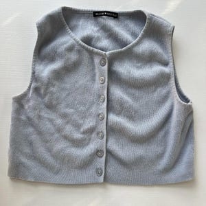 May include: A light blue, sleeveless button-up vest. The vest has a round neckline and is fastened with six small, matching buttons. The brand name "Brandy Melville" is visible on a small tag at the top. The vest is made of a soft, knitted material.
