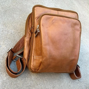 May include: A brown leather backpack with a rectangular main compartment and a smaller front pocket. The bag has adjustable shoulder straps, a top handle, and silver-toned zippers. The leather has a worn appearance.
