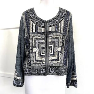 May include: A grey, black, and white beaded jacket. The jacket has a detailed geometric pattern on the front and sleeves, with a round neckline and long sleeves. It is decorated with sequins and beads, creating a textured appearance.