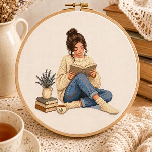 May include: A cross-stitch embroidery in a round wooden hoop featuring a person with a dark hair bun wearing a cream-colored cable knit sweater, blue jeans, and socks, sitting while reading a book next to a stack of books, a vase of lavender, and a mug.