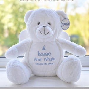 Könnte beinhalten: Ein weißer Teddybär mit gestickten Details. Der Bär hat ein lächelndes Gesicht, Engelsflügel und einen blauen gestickten Text mit der Aufschrift "Isaac Amir Whyte February 25, 2026". Der Bär hat ein Etikett mit der Aufschrift "Zippies".