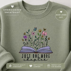 Op de afbeelding: Een olijfgroene sweater met ronde hals en een borduursel van een open boek met paarse pagina's. Kleurrijke wilde bloemen en vlinders komen uit de pagina's tevoorschijn. De tekst "JUST ONE MORE chapter" is in het zwart onder het boek geborduurd.