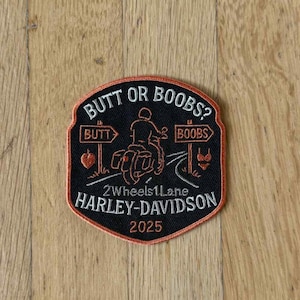 Può includere: Toppa Harley-Davidson nera e arancione con la scritta "BUTT OR BOOBS?" e "2Wheels1Lane". La toppa presenta una grafica di una moto e l'anno "2025". La toppa ha un bordo arancione.