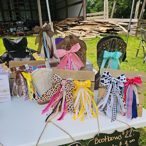 May include: A display of boot bows in various colors and patterns, including pink, leopard print, and gingham. The bows are attached to cowboy boots and displayed on a white table. A sign reads "Bootbows $22".