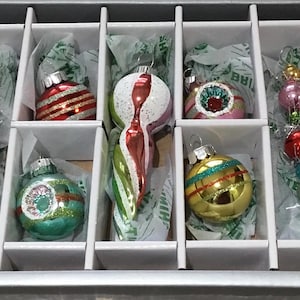 Puede incluir: Una colección de adornos navideños vintage en una caja dividida. Los adornos son de varias formas y colores, incluyendo rojo, verde, rosa y dorado. Algunos tienen rayas, purpurina o diseños intrincados. La caja es blanca con divisores.