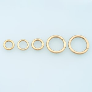 Puede incluir: Cinco anillos redondos de cierre de resorte de color dorado, de varios tamaños, están dispuestos en fila. Los cierres están abiertos, revelando el mecanismo de resorte. Los anillos son de material metálico brillante sobre fondo azul claro.